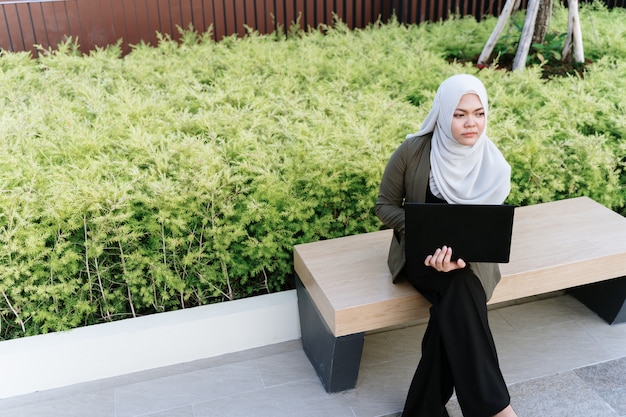 Mujer musulmana asiática joven en traje verde y trabajando en una computadora en el parque.