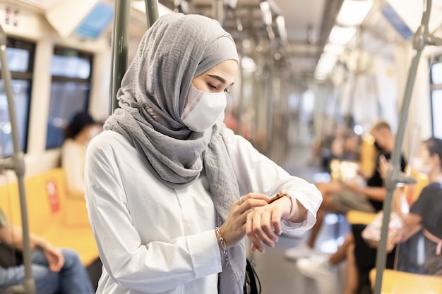 Mujer musulmana asiática con mascarilla médica para prevenir el polvo y el virus de la infección y mirando smartwatch en el sistema de transporte público Skytrain