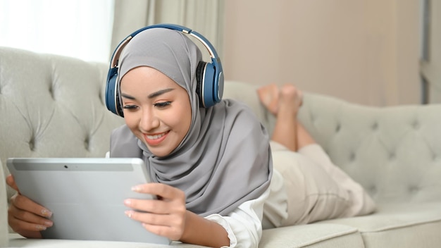 Mujer musulmana asiática relajada recostada en un sofá escuchando música con auriculares y usando una tableta