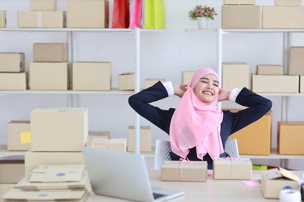 Mujer musulmana asiática religiosa en traje azul y eje rosa sentado con emoción de felicidad y relajación. La mujer de negocios se sienta con el fondo de la entrega de la caja de la PYME del paquete. Concepto de trabajo en casa