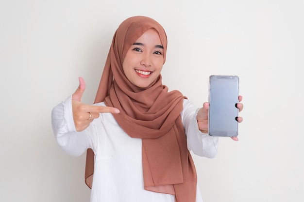 Foto mujer musulmana asiática sonriendo y señalando a la pantalla en blanco del teléfono móvil que sostiene