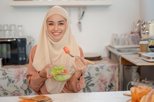 Una mujer musulmana asiática sonriente comiendo un plato de ensalada saludable en una mesa en la cocina