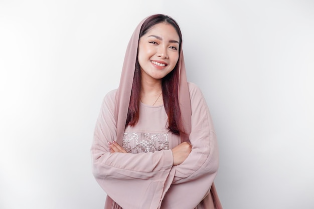 Una mujer musulmana asiática sonriente y confiada que lleva un pañuelo en la cabeza de pie con los brazos cruzados y mirando la cámara aislada sobre fondo blanco