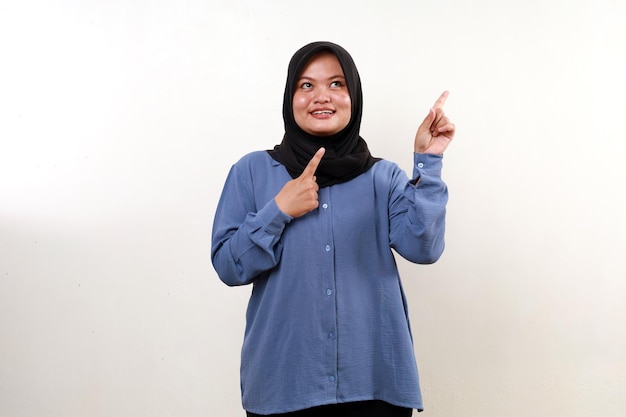 Una mujer musulmana asiática sonriente de pie mientras señala hacia arriba aislada en blanco
