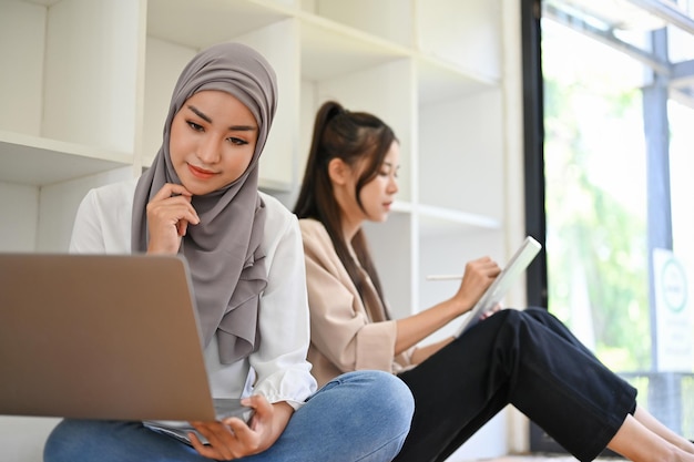 Mujer musulmana asiática usa una computadora portátil para administrar su tarea en la biblioteca con su amiga