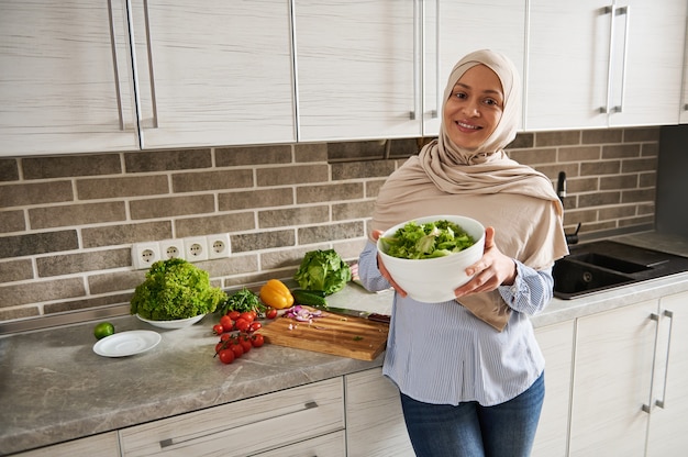 Mujer musulmana con la cabeza cubierta en hijab sostiene un cuenco con ensalada y sonríe a la cámara de pie en una cocina doméstica con verduras sobre una mesa