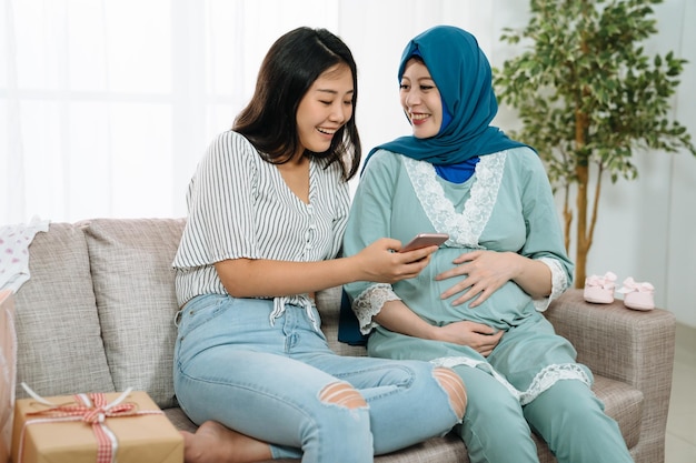 Mujer musulmana embarazada y amiga de la dama usando compras de teléfonos móviles en línea en la venta de verano juntos. niña feliz con la maternidad celebrando la fiesta de la ducha del bebé con cajas de regalo en el sofá en la sala de estar de la casa.