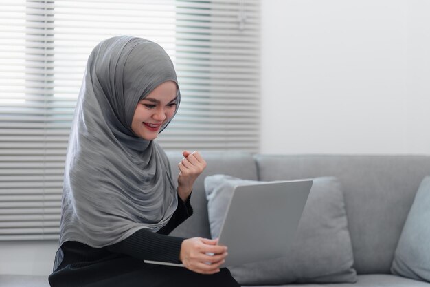 Mujer musulmana emocionada con un hijab mostrando un gesto de victoria mientras mira la pantalla de su portátil