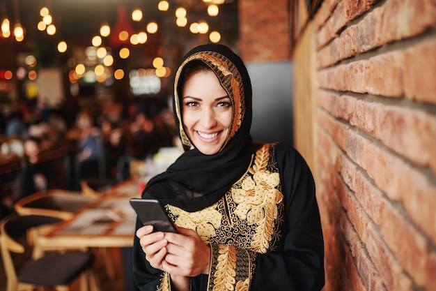 Mujer musulmana encantadora alegre en abaya usando el teléfono inteligente para leer o escribir mensajes mientras está sentado en la cafetería.