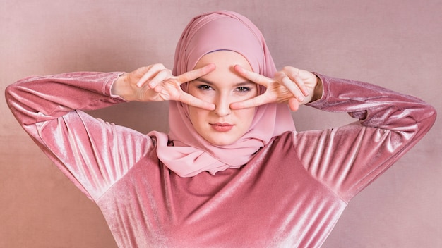 Mujer musulmana enojada que muestra v muestra cerca de sus ojos sobre superficie coloreada