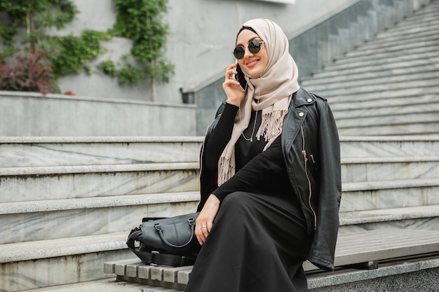 Mujer musulmana con estilo moderno en hijab, chaqueta de cuero y abaya negra caminando en las calles de la ciudad hablando por teléfono inteligente