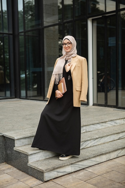 Mujer musulmana con estilo moderno en hijab, chaqueta de estilo empresarial y abaya negra caminando en las calles de la ciudad con laptop