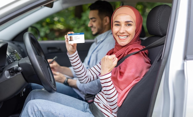 Mujer musulmana feliz en hiyab mostrar licencia de conducir y puño en puerta abierta en auto después de examen con hombre