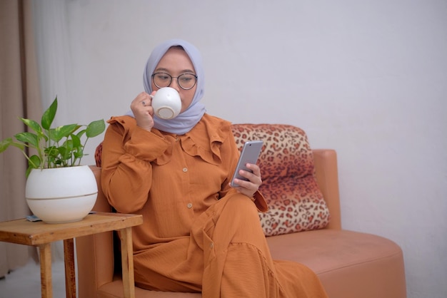 Mujer musulmana feliz relajarse en casa mientras toma té y mira la pantalla del teléfono