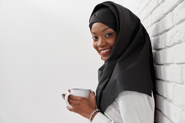 Mujer musulmana en hijab apoyada en la pared sosteniendo una taza blanca