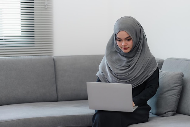 Mujer musulmana con hijab concentrándose en su trabajo en una computadora portátil mientras está sentada en un sofá en una casa