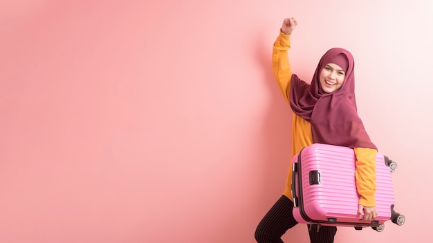 Mujer musulmana con hijab sostiene equipaje sobre fondo rosa, concepto de viaje de personas