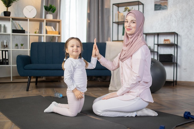 Mujer musulmana con hiyab sentada de rodillas en una alfombra y choca los cinco con su pequeña hija
