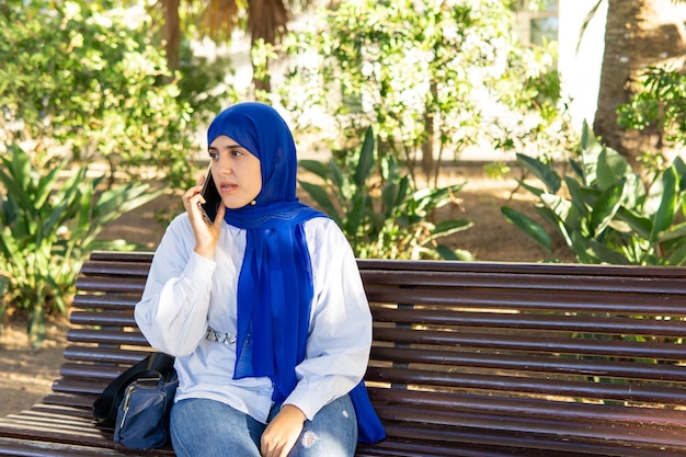 Foto mujer musulmana joven con hijab sentada en un banco del parque hablando por teléfono