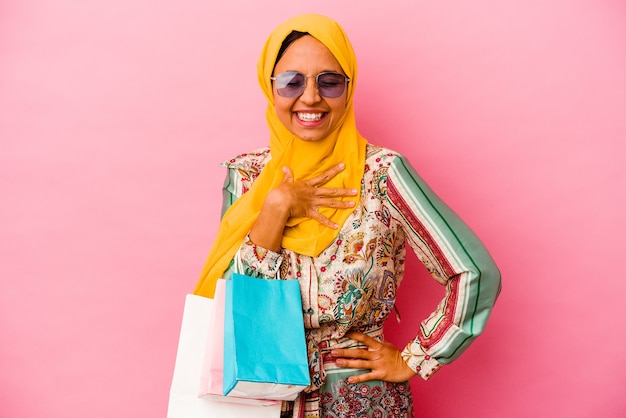Mujer musulmana joven que compra ropa aislada en la pared rosada se ríe a carcajadas manteniendo la mano en el pecho.