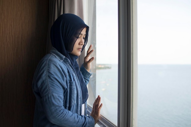 Mujer musulmana joven y triste asiática frente a una gran ventana de vidrio mirando la pacífica vista del océano ambiente de mal humor