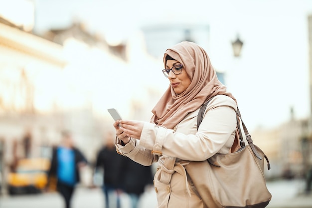 Mujer musulmana de mediana edad que usa hijab con una cara feliz parada en un entorno urbano, enviando mensajes en su teléfono inteligente.