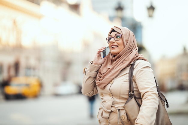 Mujer musulmana de mediana edad que usa hijab con una cara feliz parada en un entorno urbano, tomando su teléfono inteligente.