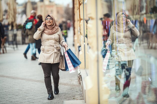 Una mujer musulmana de mediana edad que usa hiyab con una cara feliz camina en un entorno urbano, va de compras con bolsas de papel de colores y habla por teléfono inteligente.