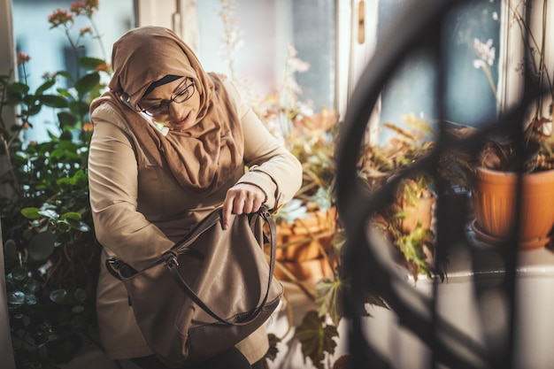 Una mujer musulmana de mediana edad que usa hiyab con una cara feliz está parada junto a la ventana buscando algo en su bolso y tomando su teléfono inteligente.