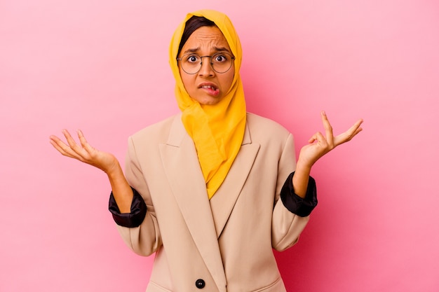 Mujer musulmana de negocios joven aislada sobre fondo rosa confundido y dudoso encogiéndose de hombros para mantener un espacio de copia.