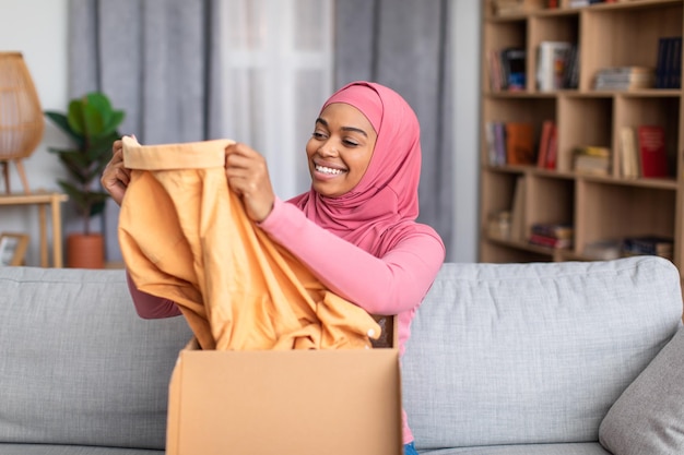 Mujer musulmana negra feliz abriendo caja de cartón revisando su entrega sosteniendo ropa nueva y sonriendo