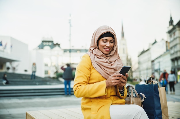 Mujer musulmana negra feliz usando un hiyab y enviando mensajes en su teléfono inteligente mientras está sentada en el centro.