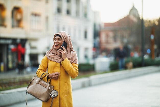 Mujer musulmana negra sonriente usando un hijab y hablando en su teléfono inteligente mientras camina en un entorno urbano.