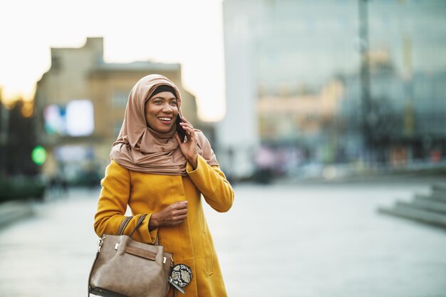 Mujer musulmana negra usando hiyab con una cara feliz parada en un entorno urbano y usando su teléfono inteligente.