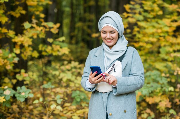 Mujer musulmana en el parque con teléfono inteligente conectado de forma inalámbrica en línea. Espacio para publicidad