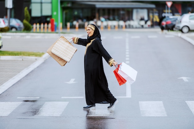 Mujer musulmana sonriente positiva llena de alegría con ropa tradicional que lleva bolsas de compras en las manos y se siente satisfecha con sus compras mientras cruza la calle.