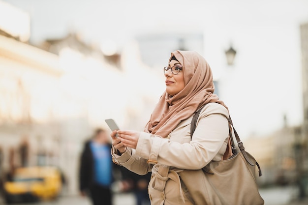 Mujer musulmana sonriente usando un hiyab y usando su teléfono inteligente mientras está de pie en un entorno urbano.