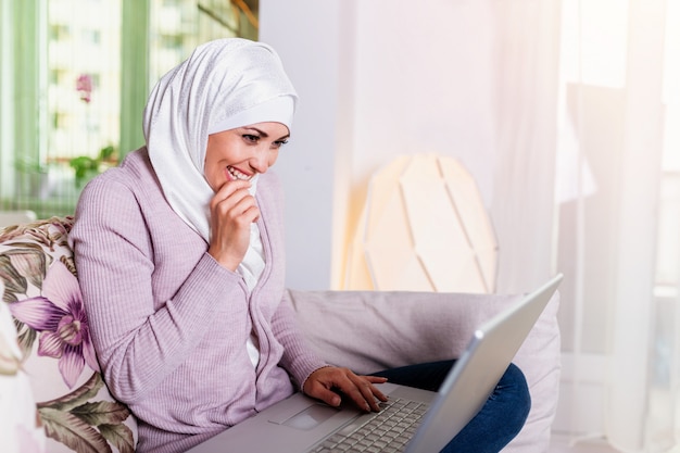 Mujer musulmana usando su computadora portátil, navegando por las redes sociales en casa