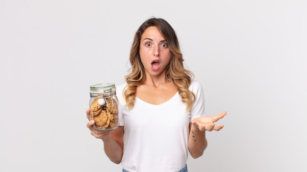 Mujer muy delgada asombrada, conmocionada y asombrada con una sorpresa increíble y sosteniendo una botella de vidrio de galletas