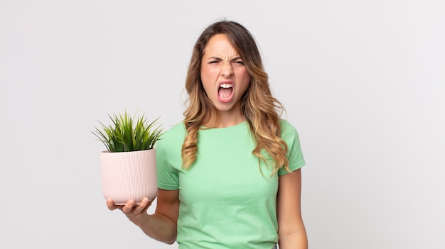 Mujer muy delgada gritando agresivamente, luciendo muy enojada y sosteniendo una planta decorativa