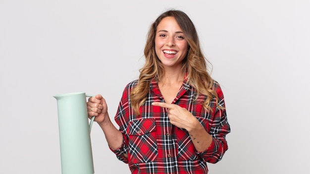 Mujer muy delgada mirando emocionada y sorprendida apuntando hacia un lado y sosteniendo un termo de café