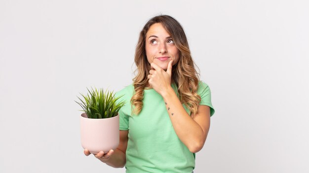 Mujer muy delgada pensando, sintiéndose dudosa y confundida y sosteniendo una planta decorativa