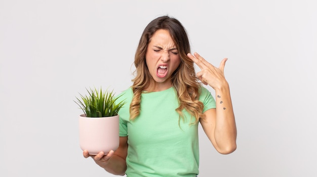 Mujer muy delgada que parece infeliz y estresada, gesto de suicidio haciendo un signo de pistola y sosteniendo una planta decorativa