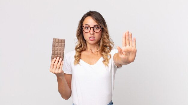 Mujer muy delgada que parece seria mostrando la palma abierta haciendo gesto de parada y sosteniendo una barra de chocolate