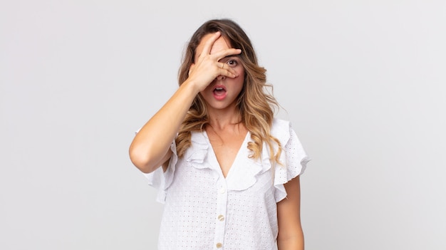 Mujer muy delgada que parece sorprendida, asustada o aterrorizada, cubriéndose la cara con la mano y mirando entre los dedos