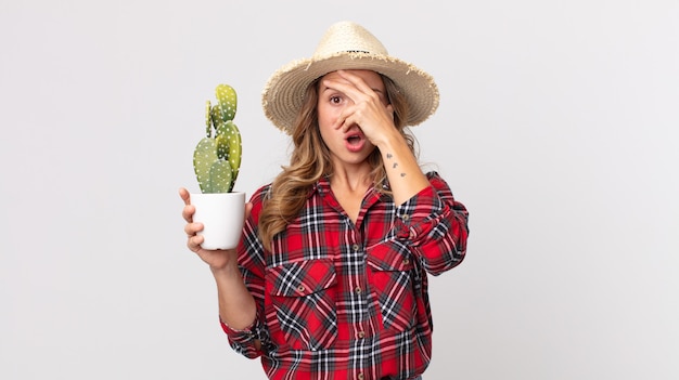 Mujer muy delgada que parece sorprendida, asustada o aterrorizada, cubriéndose el rostro con la mano sosteniendo un cactus. concepto de granjero