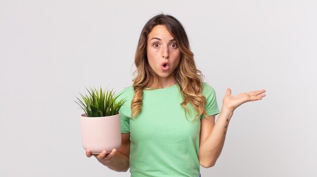 Mujer muy delgada que parece sorprendida y conmocionada, con la mandíbula caída sosteniendo un objeto y sosteniendo una planta decorativa