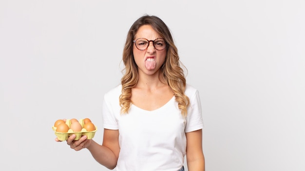 Mujer muy delgada que se siente disgustada e irritada y con la lengua fuera y sosteniendo una caja de huevos