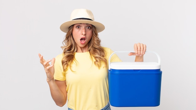 Mujer muy delgada que se siente extremadamente conmocionada y sorprendida y sosteniendo una nevera de picnic de verano