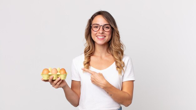 Mujer muy delgada sonriendo alegremente, sintiéndose feliz y apuntando hacia un lado y sosteniendo una caja de huevos